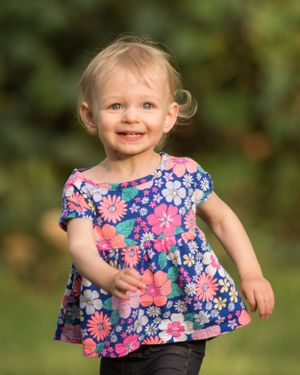 A small child wearing a colorful floral dress runs and smiles outdoors against a blurred green background.