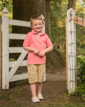 A child in a coral polo shirt and khaki shorts stands smiling by a white garden gate and rustic fence in a wooded setting.