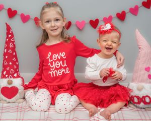 Two children wearing red and white Valentine's Day themed outfits pose together against a backdrop with heart decorations.