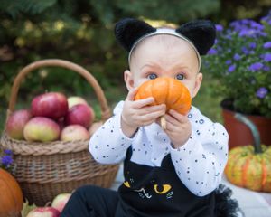 Small child in black cat overalls holding an orange pumpkin, with autumn harvest basket and decorations in outdoor garden.