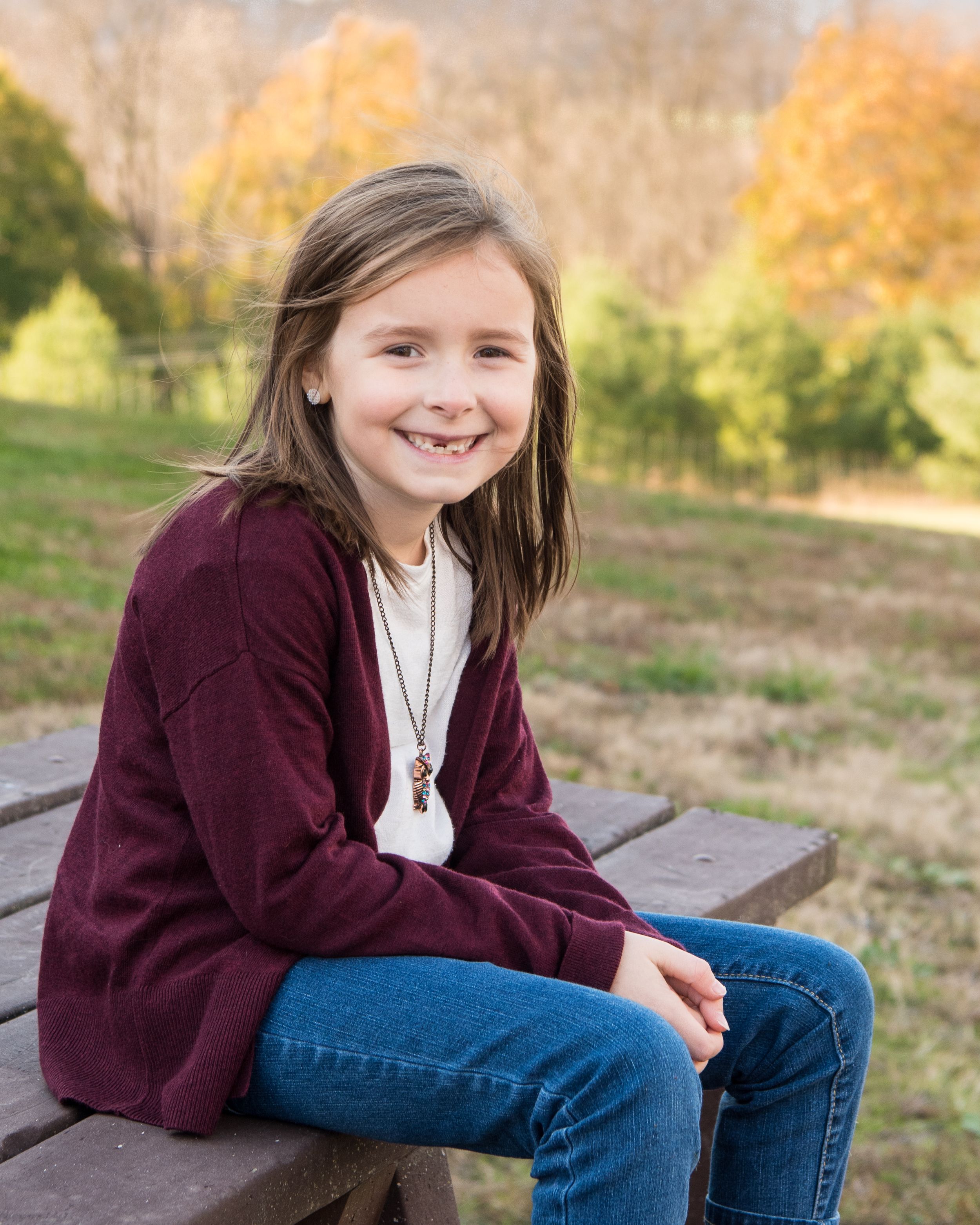 Young person in a burgundy cardigan and jeans sits on a picnic table outdoors with autumn foliage in the background.