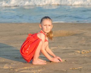 Someone in a bright red dress sits thoughtfully on the sandy beach at sunset as waves roll in behind them.