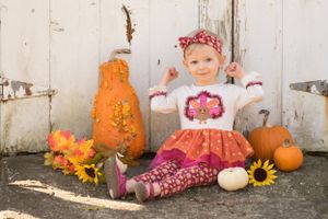 A small girl sitting in a fall harvest display with decorated pumpkins, sunflowers, and small gourds against rustic white wooden backdrop.