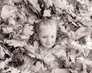Black and white image of a small child lying in a pile of autumn leaves while smiling at the camera.