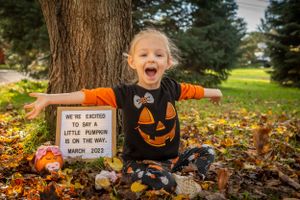 A child in a pumpkin shirt sits on fall leaves next to a Halloween sign and decorative pumpkins in an outdoor setting.