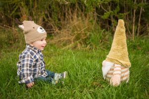 Child in plaid shirt and hat sitting in grass facing a large plush garden gnome decoration.