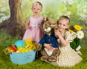 Studio portrait of two young children posing with Easter decorations and a stuffed bunny against a spring garden backdrop.