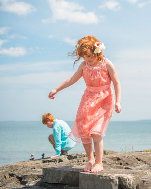 Two children explore rocky coastal outcrop on sunny day, with one wearing coral dress and other in a turquoise shirt by the ocean.