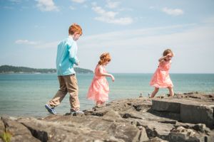 Three children in colorful clothes walk along rocky coastal cliffs overlooking a serene blue ocean on a sunny day.