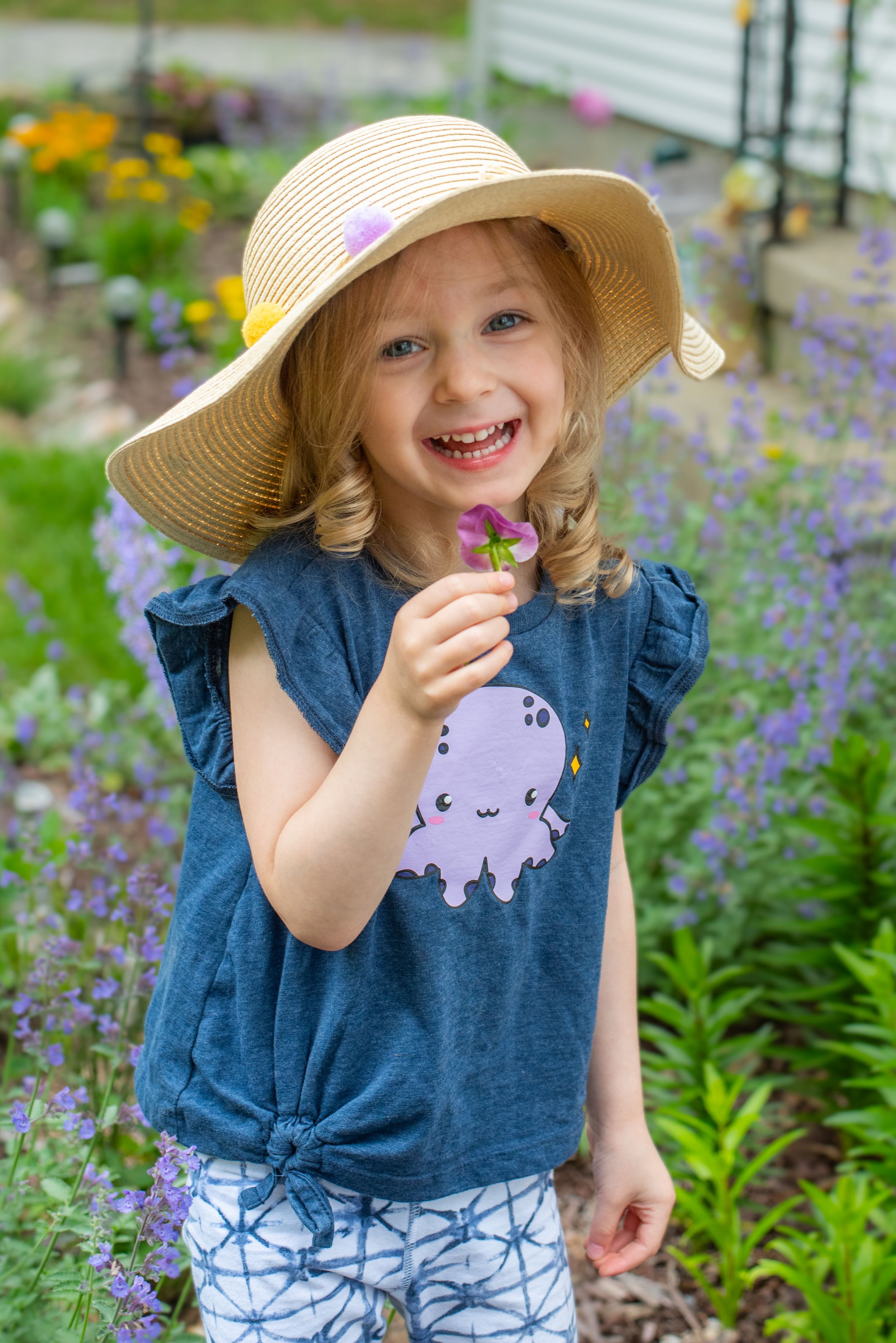 Young child in a straw hat and blue shirt with octopus design poses joyfully among blooming purple flowers in a garden.
