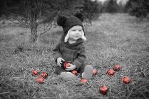 A small child in winter clothing sits in a field with red Christmas ornaments scattered around in black and white photography.