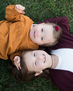 Two children laying on green grass wearing autumn colored clothing and smiling at the camera.