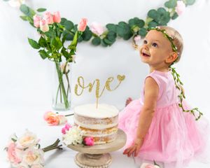 First birthday celebration setup for a little girl in a pink dress, white cake, floral decorations and gold 'one' cake topper.