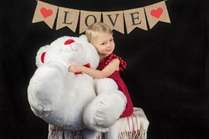 A small figure in a red dress sits on a chair hugging a large white teddy bear beneath a rustic LOVE banner.