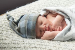 Sleeping newborn baby wrapped in soft gray blanket wearing knit hat resting on textured white surface.