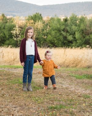 Two siblings in casual autumn outfits walk together in a field with mountains and trees in the background.