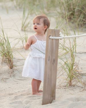 A small child in a white sundress stands next to a wooden post on a sandy beach with sea grass in the background.