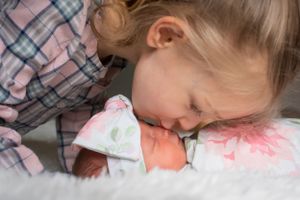 A tender moment between siblings as an older child gently kisses a newborn baby swaddled in white.