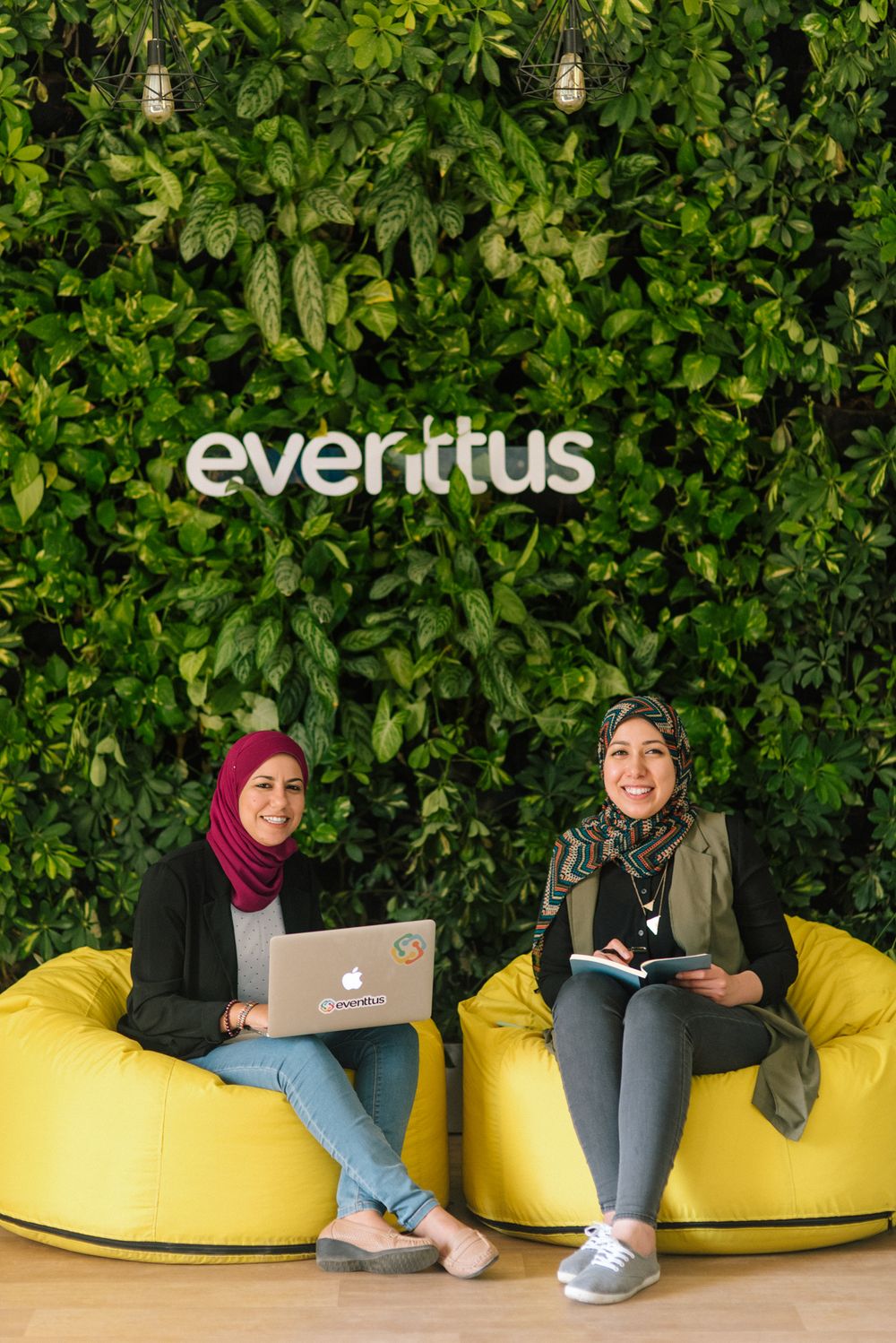 Two colleagues working on laptops while sitting on yellow bean bags against leafy Everitus wall.