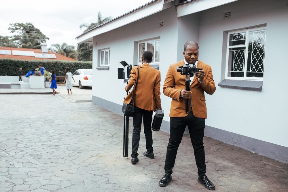Two photographers in matching brown jackets checking their camera equipment outside a building.
