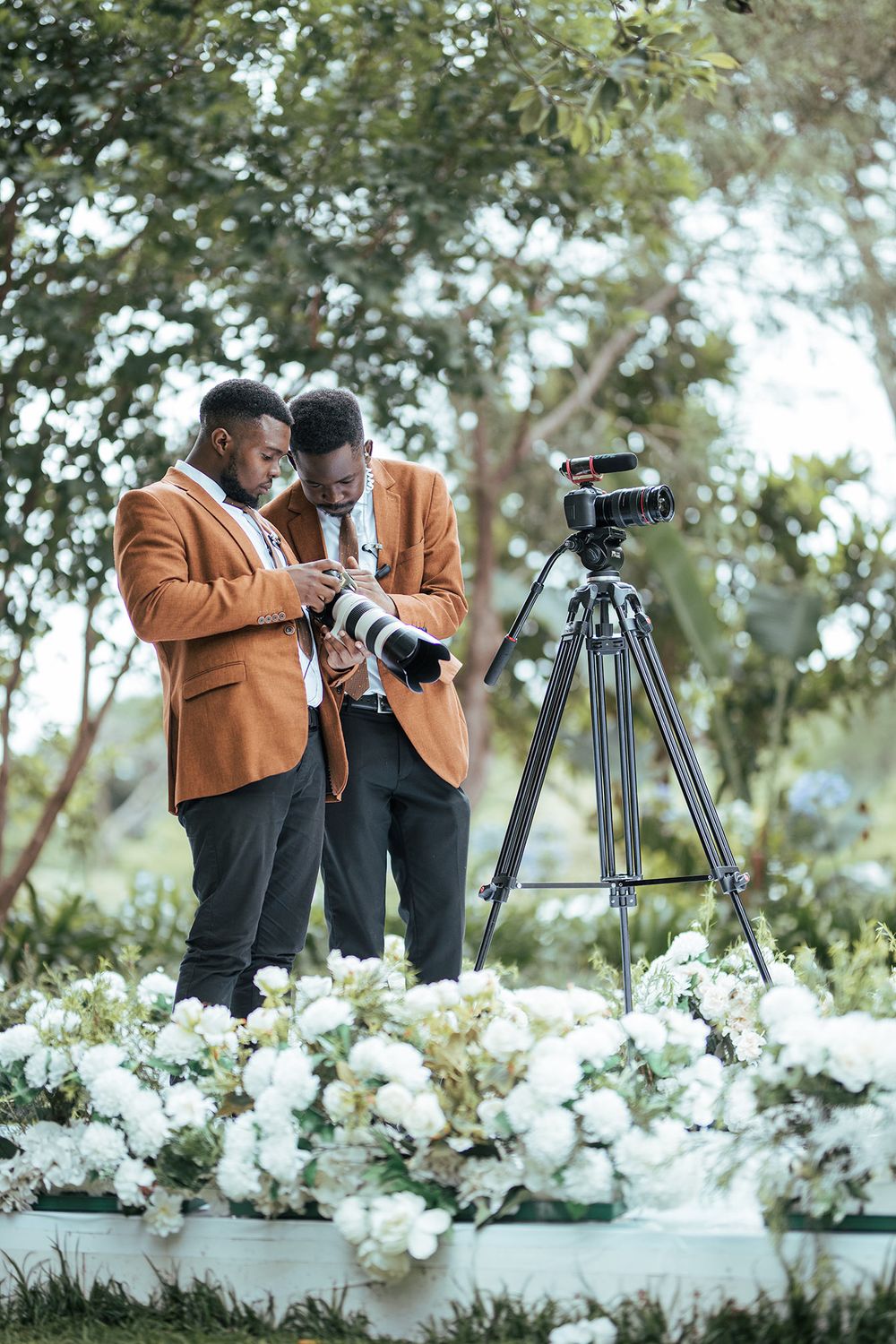 Two photographers in matching blazers reviewing images near white floral arrangements and camera setup.