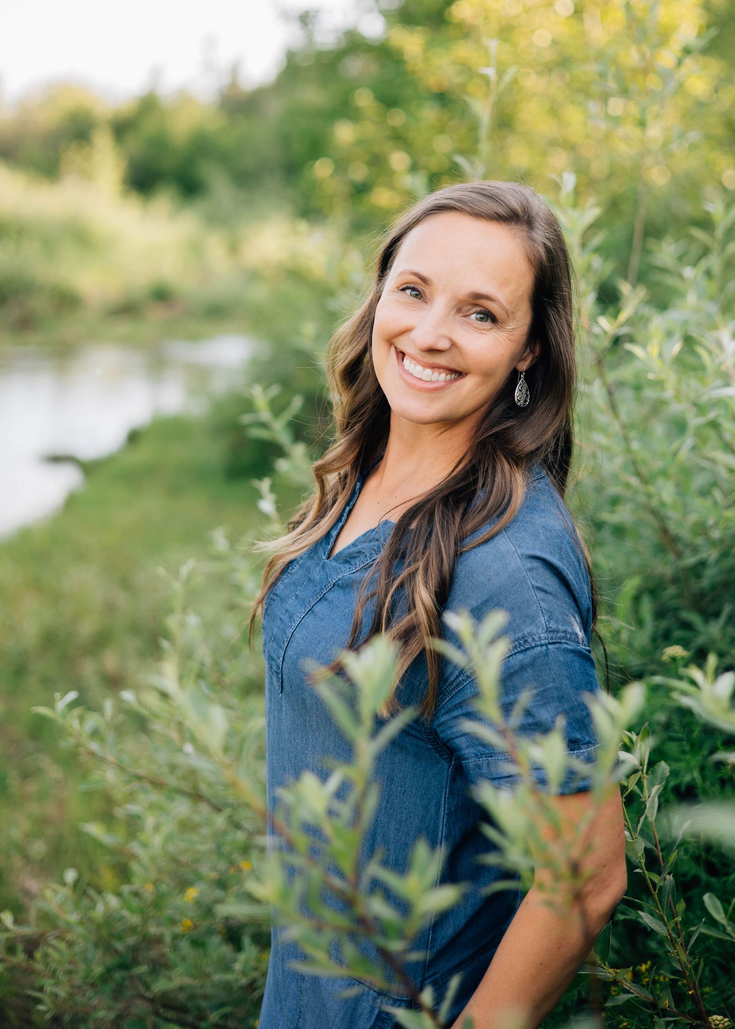 Woman in denim shirt smiling in natural outdoor setting with green foliage.