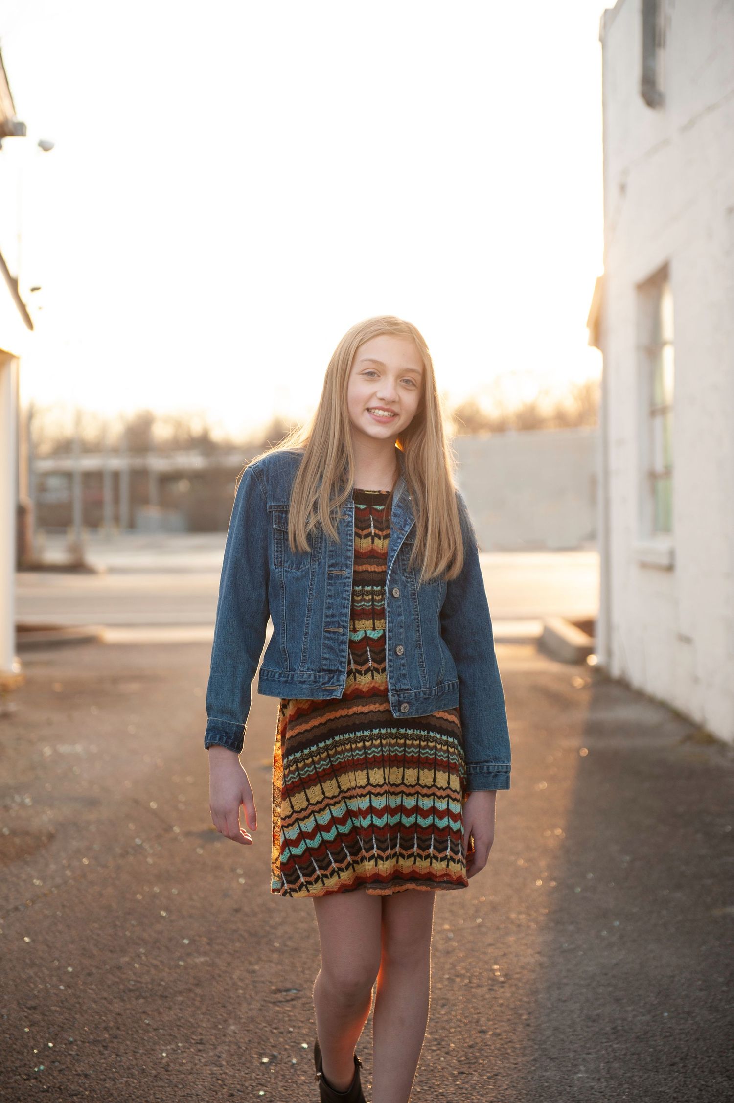 Model in denim jacket and patterned dress poses in golden sunset light.