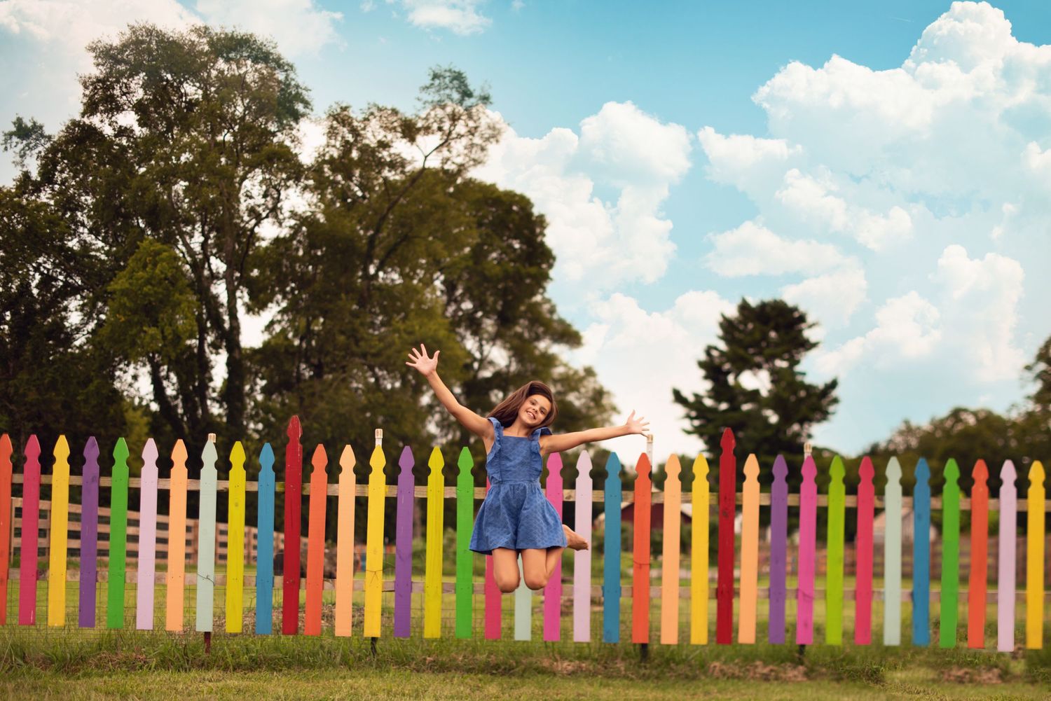 Someone jumps playfully in front of a vibrant rainbow-colored picket fence on a sunny day.