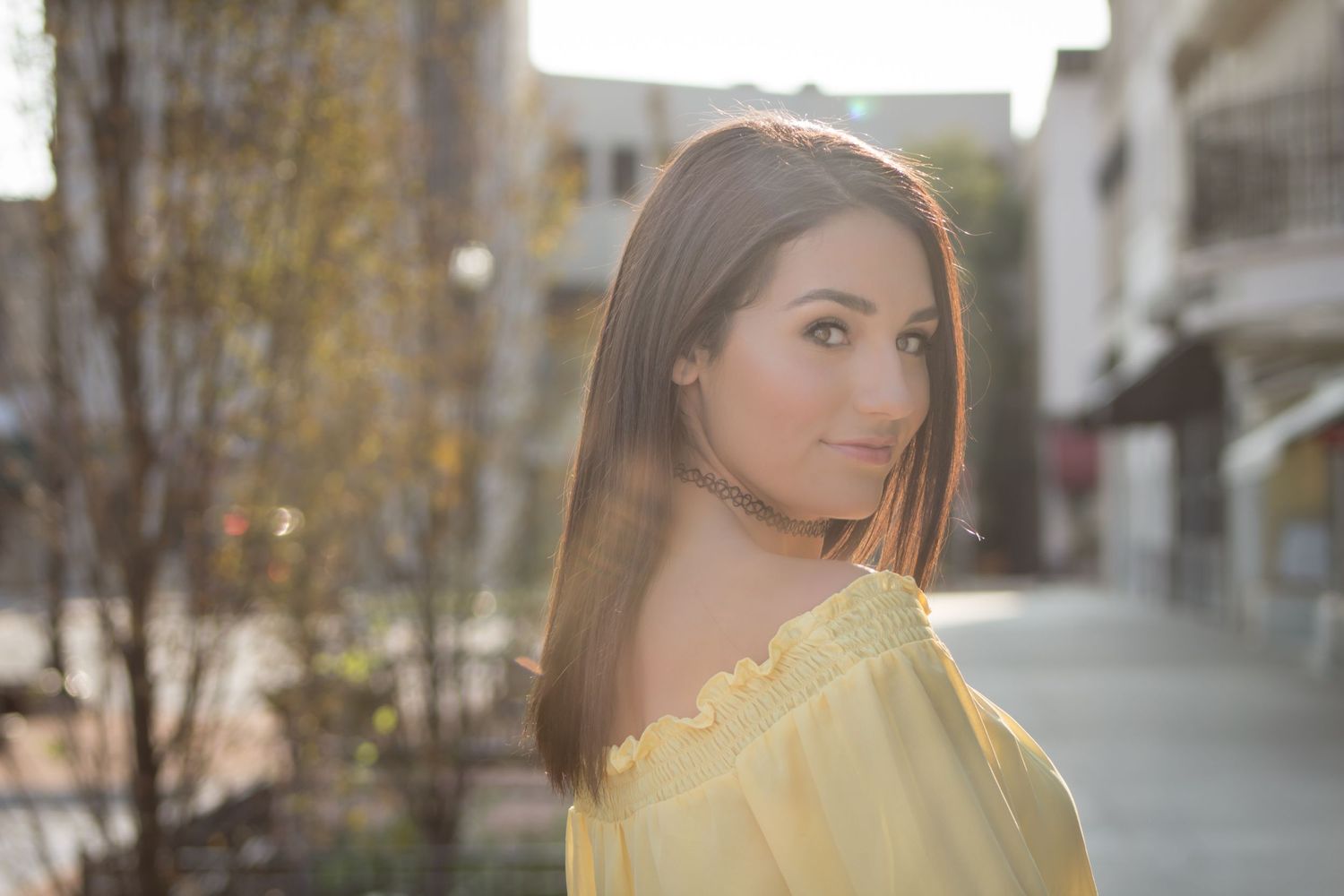 Young woman in yellow off-shoulder top poses outdoors in golden sunlight with soft bokeh background.