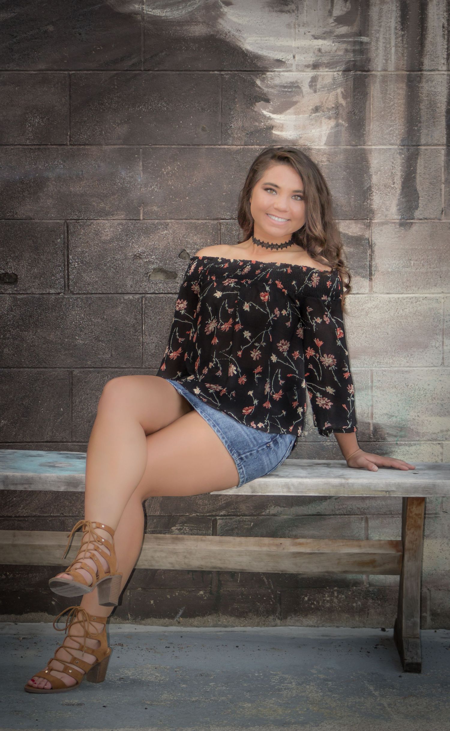 Brunette woman in floral off-shoulder top and denim shorts poses on bench with heeled sandals.