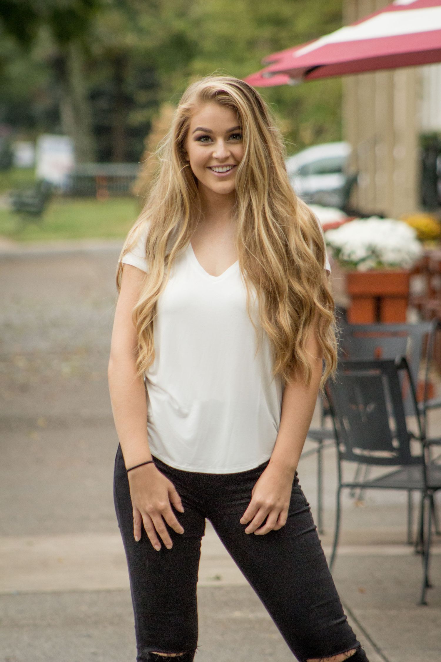 Blonde woman with long wavy hair smiles wearing white top and black ripped jeans outdoors.