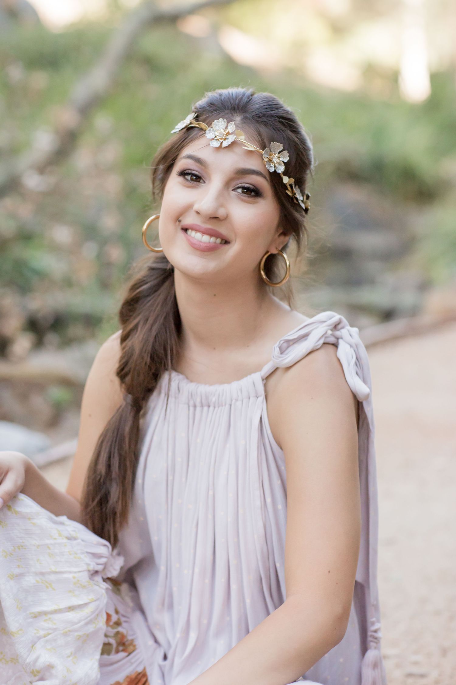 Smiling young woman wearing floral gold crown and lavender dress poses outdoors in natural setting.