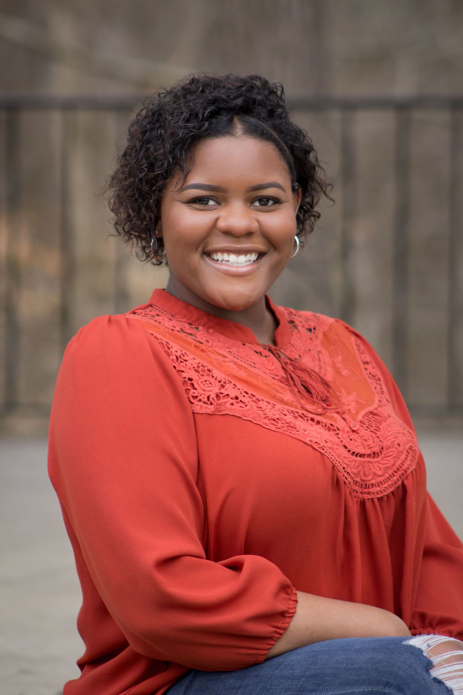 Smiling young woman in a red lace blouse sitting outdoors against a stone wall background.