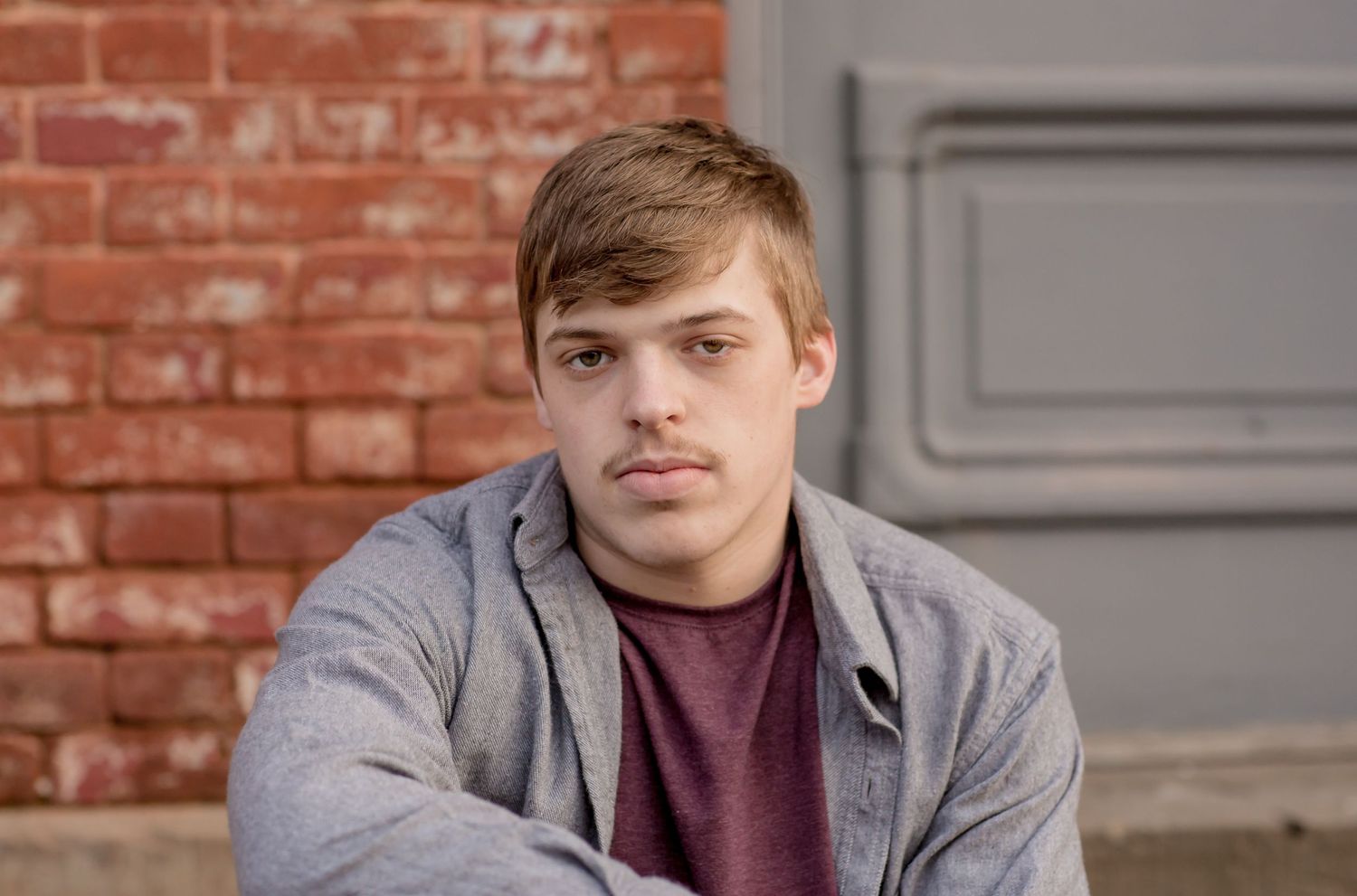 Young man in a gray hoodie with arms crossed sitting against a red brick wall.