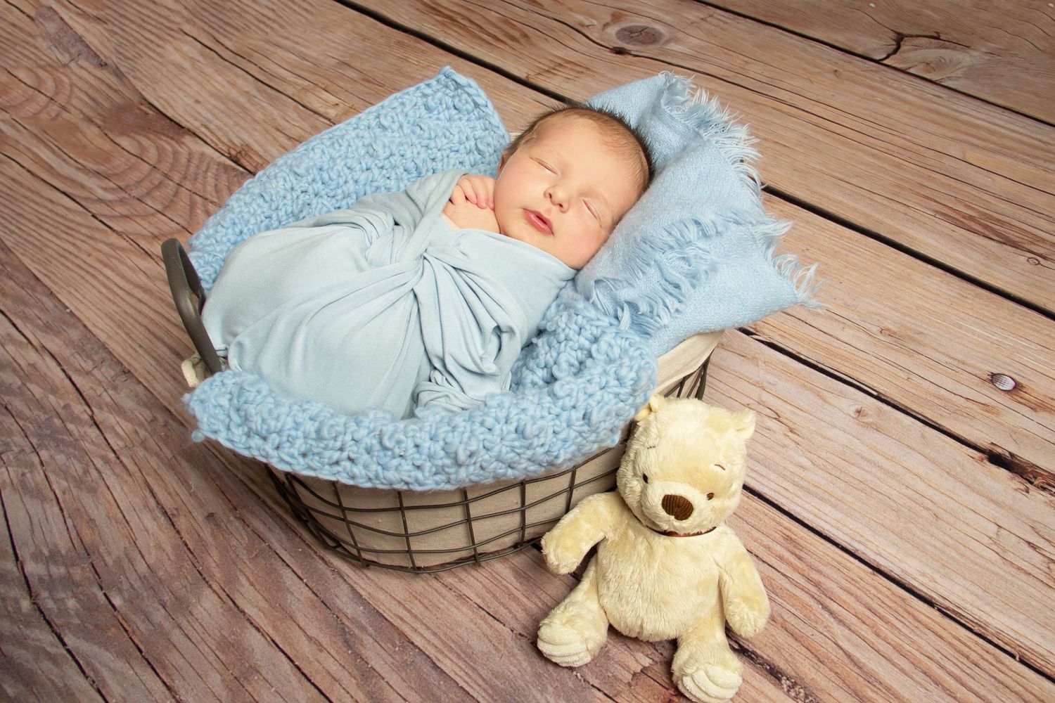 newborn baby sleeping in a wire basket wrapped in blue with a teddy