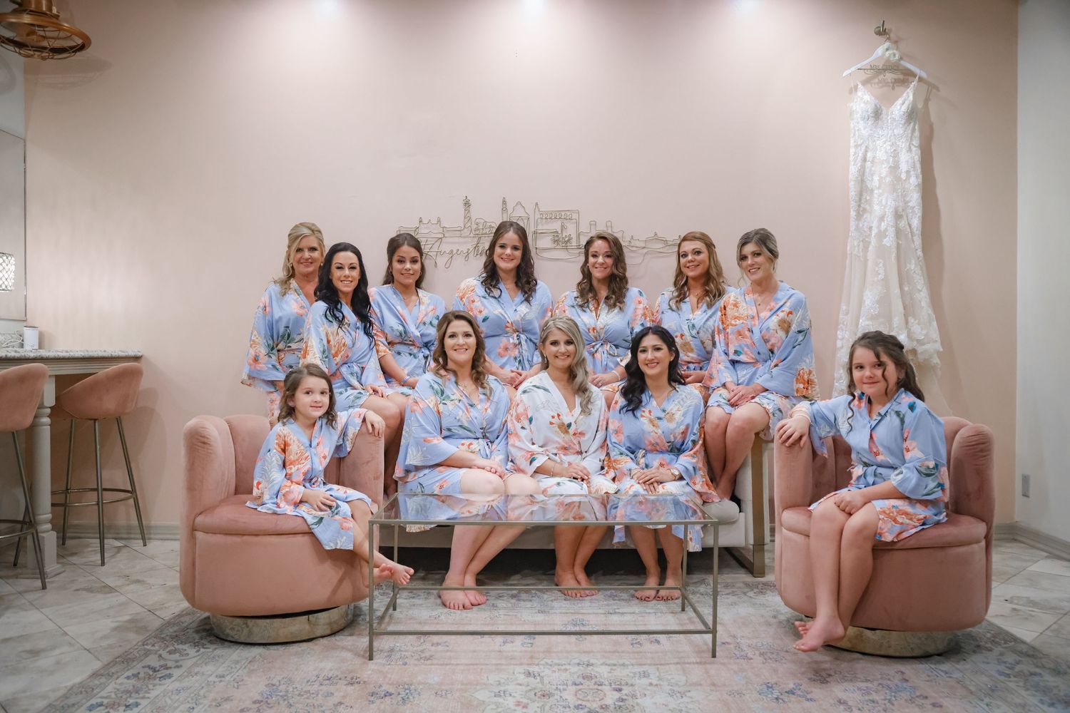 A group in matching floral robes pose together on pink furniture with a wedding dress hanging in the background.