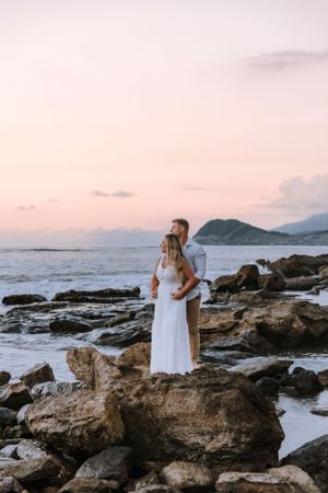 Couple in white clothing poses on rocky coastline during sunset.