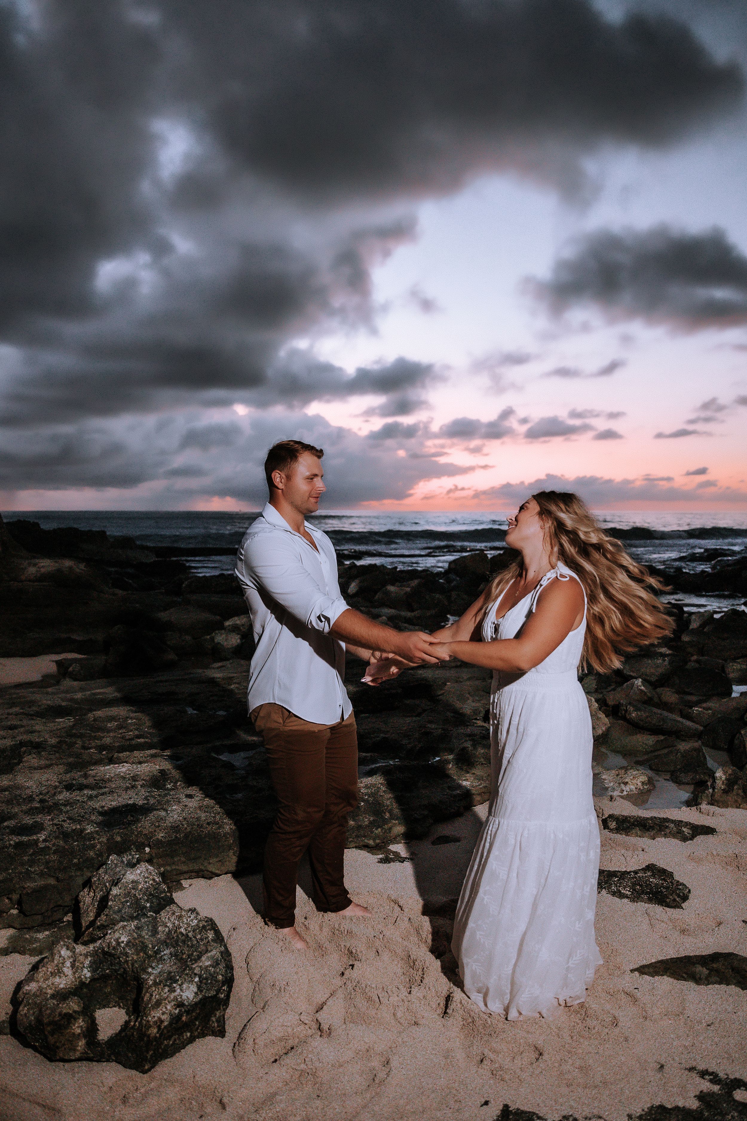 Couple in white clothing dancing and laughing on a beach with dramatic stormy clouds at sunset.