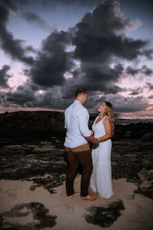 Couple in white and brown attire sharing an intimate moment on rocky coastline under dramatic sunset sky.