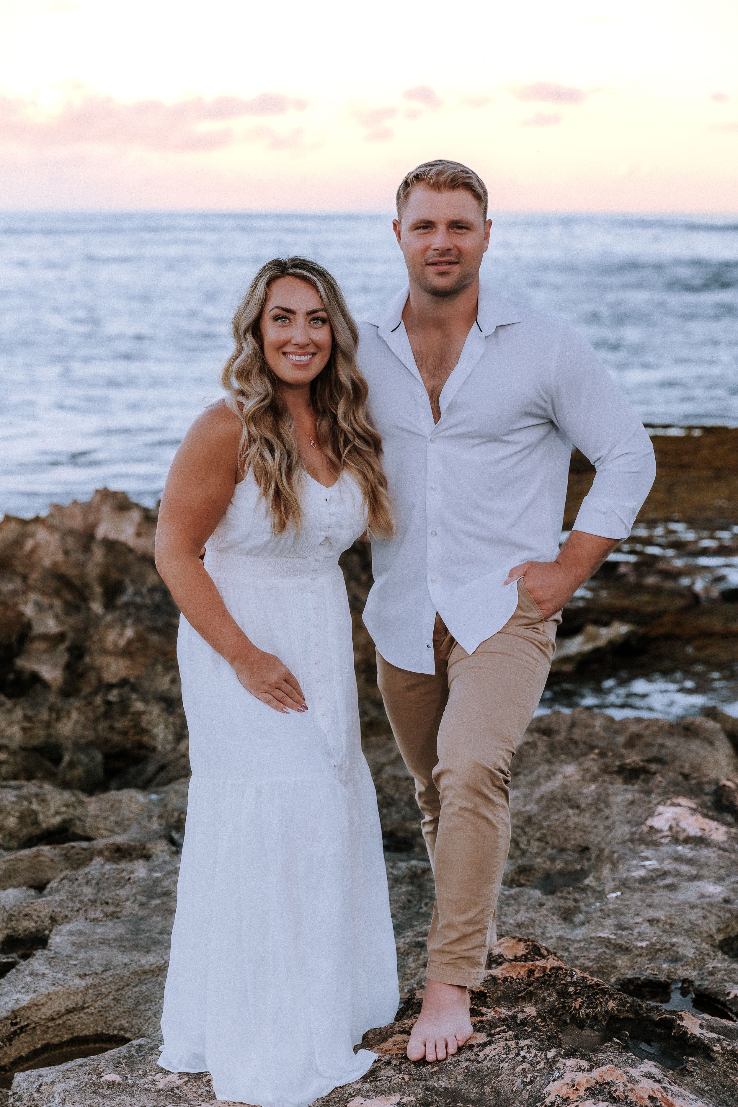 Couple poses on rocky beach at sunset wearing coordinating white and beige outfits.