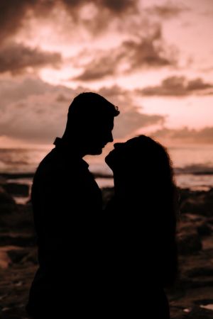 Silhouetted couple against dramatic sunset sky on rocky beach.