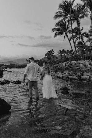 Black and white photo of couple walking through shallow ocean water near palm trees.