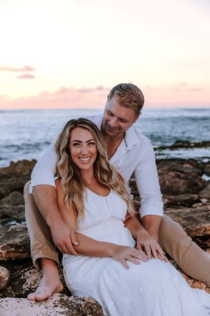 Couple relaxes on coastal rocks during sunset in matching white outfits.
