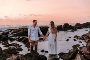 Couple walks hand in hand along rocky beach during pink sunset.