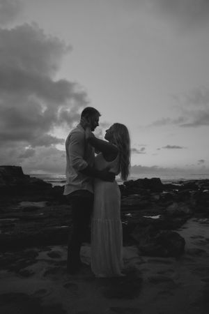 A romantic black and white silhouette of a couple embracing on a rocky beach at sunset.