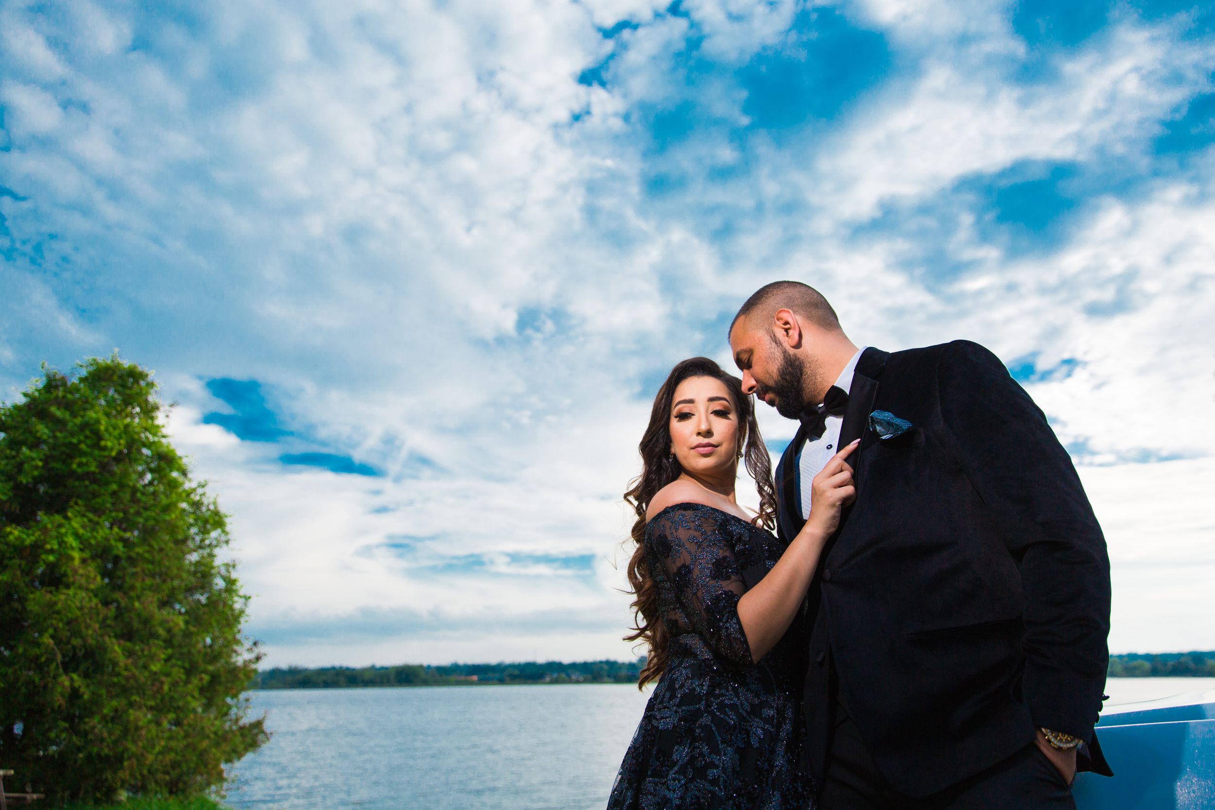 Romantic couple poses by white sports car under dramatic blue sky.