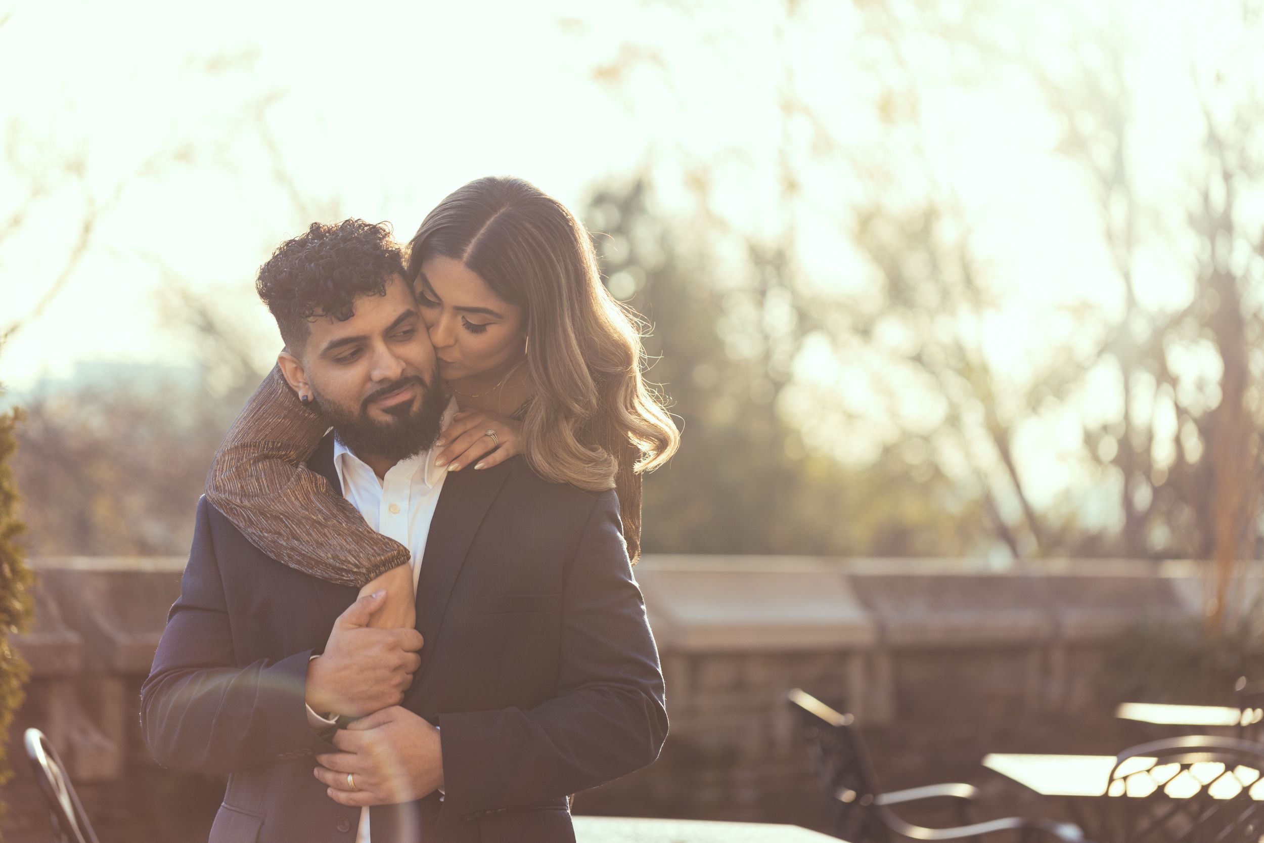 A romantic couple embraces outdoors during golden hour with warm sunlit bokeh in the background.