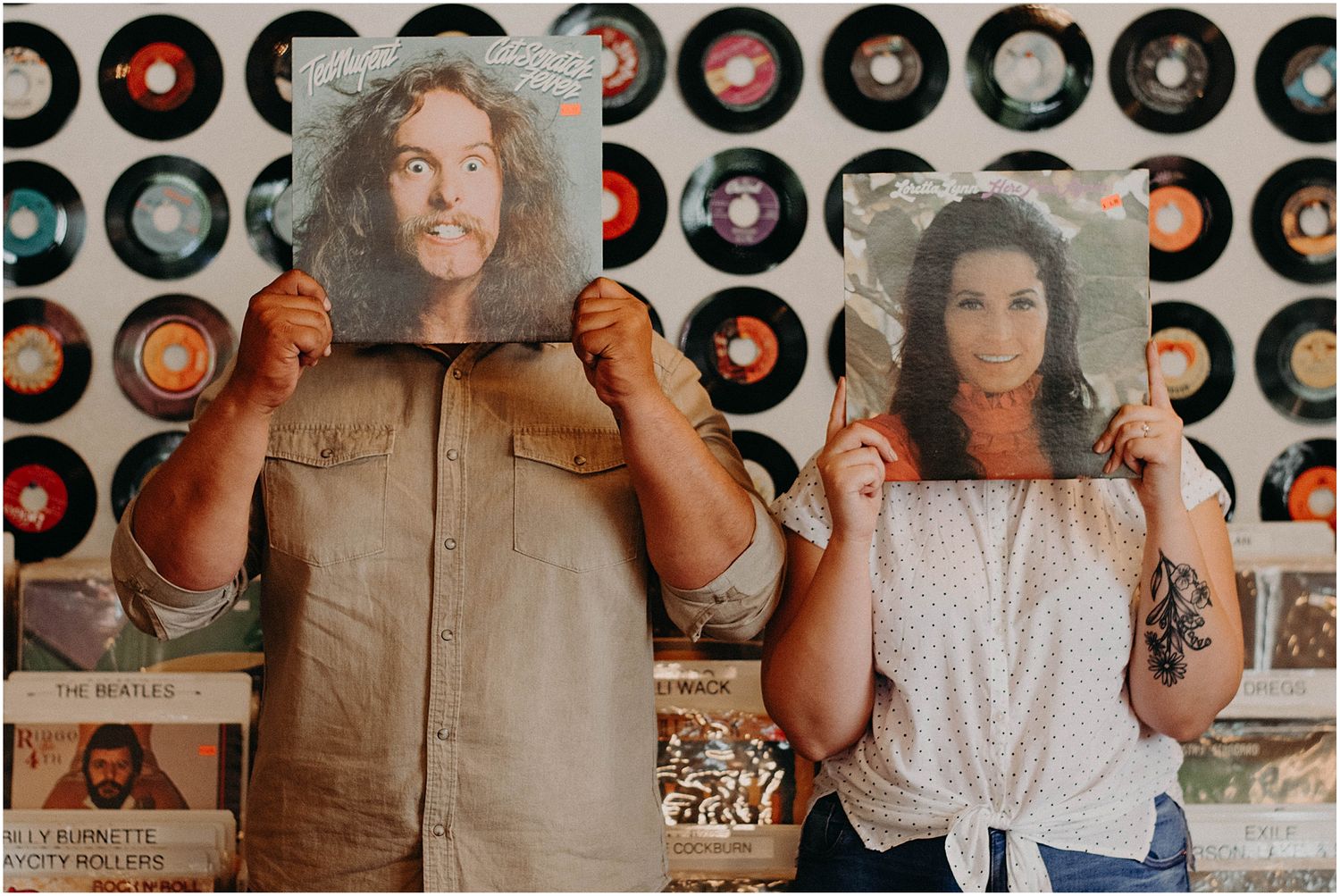 Two people posing with vinyl records covering their faces against a wall of vintage albums.