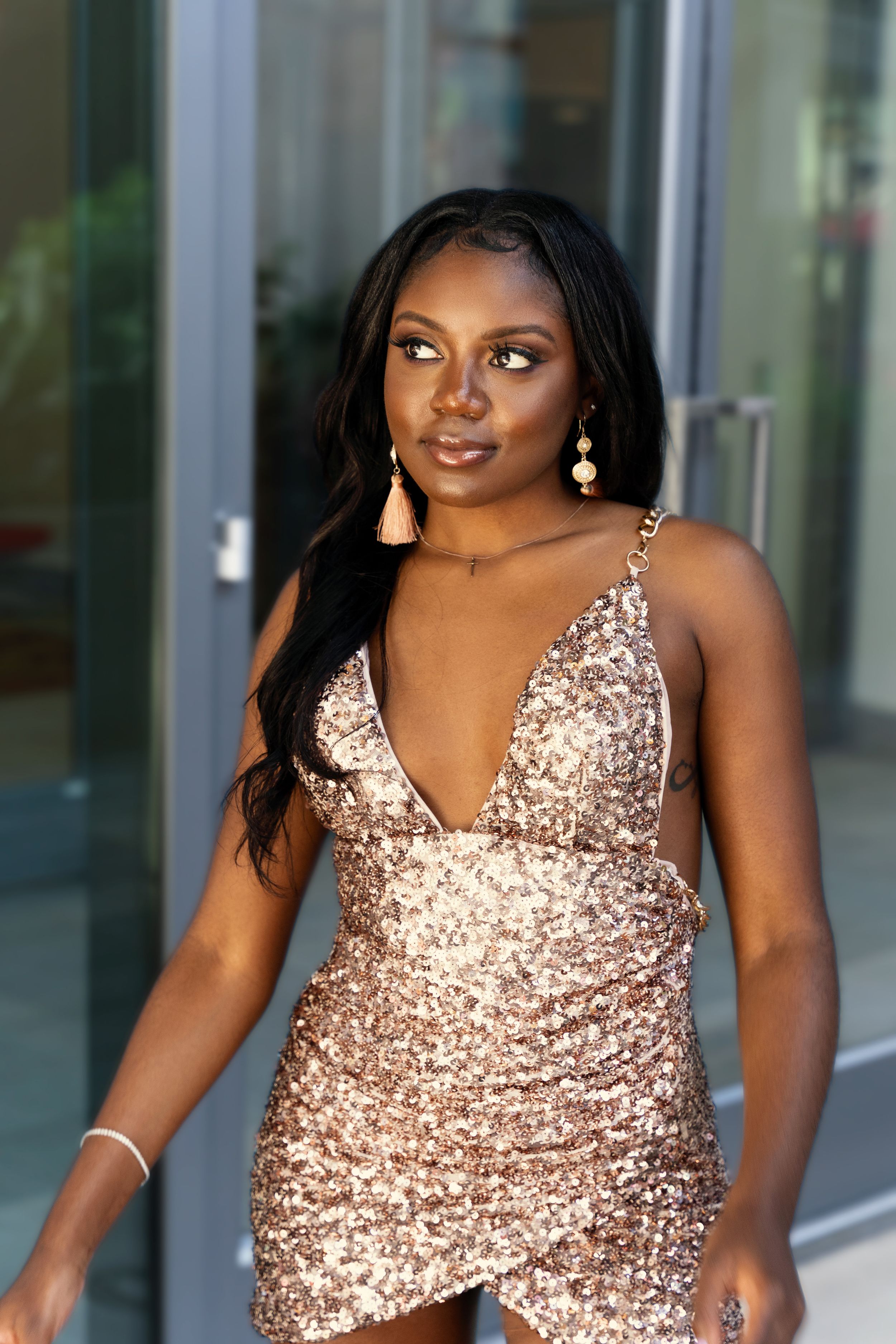 Elegant poses in rose gold sequined dress against modern glass building facade.