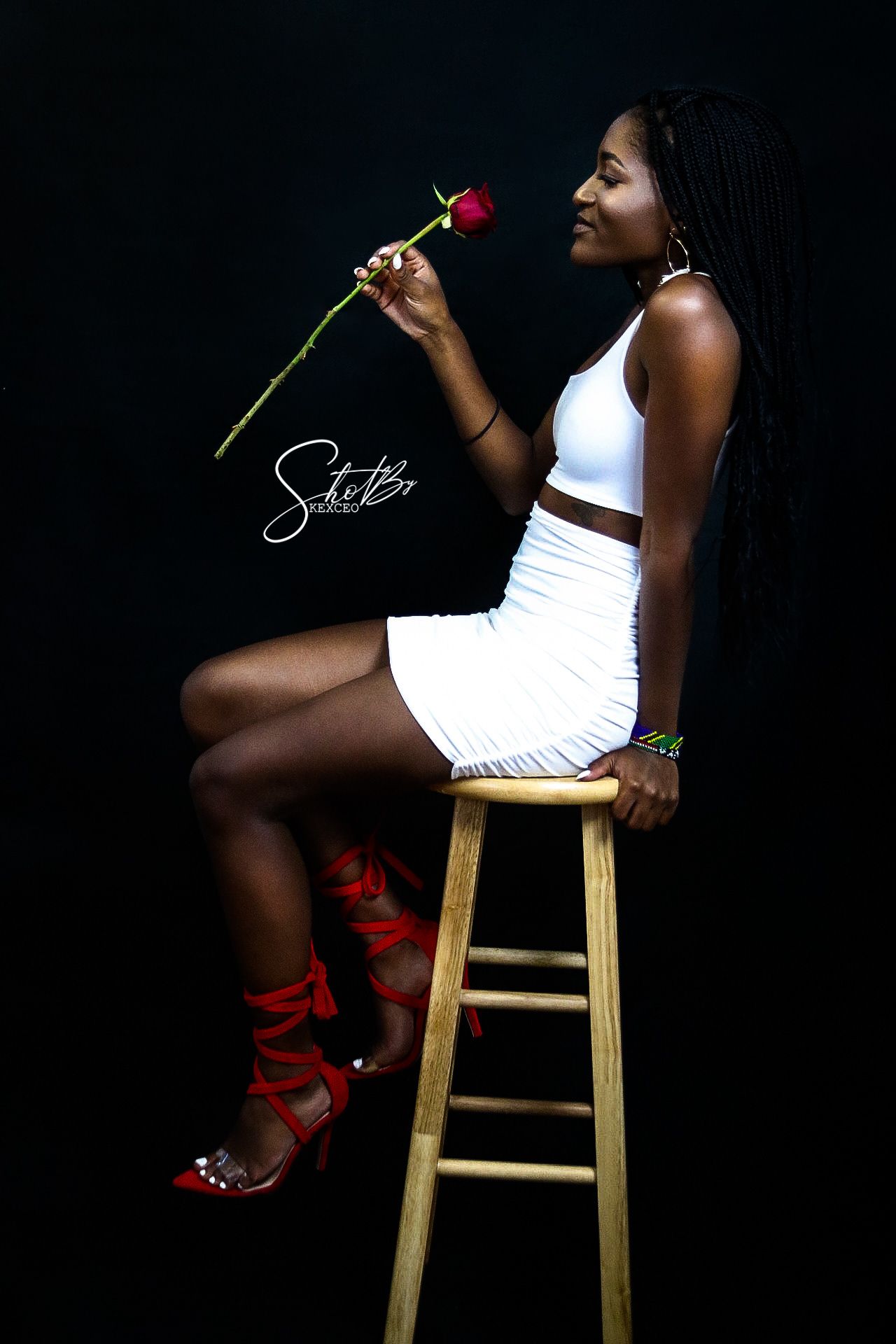 Portrait of a model in white dress holding a red rose while seated on a wooden stool.